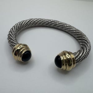May include: A silver-tone cuff bracelet featuring a twisted cable texture throughout the band. The two open ends are finished with polished gold-tone metal caps, each set with a smooth, round, black stone cabochon at the center.