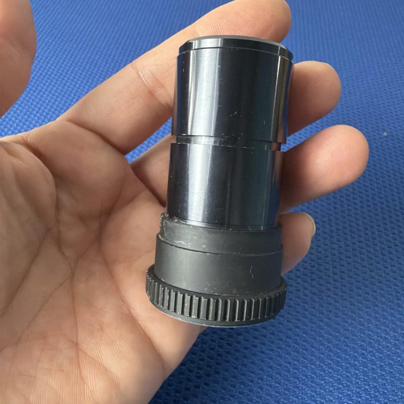 May include: A black telescope eyepiece with a rubber grip and a toothed base. The eyepiece is cylindrical and has a glossy finish. The image is taken against a blue background.