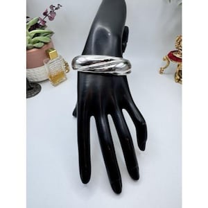 May include: A silver-toned bangle bracelet with a textured, wave-like design. The wide cuff bracelet is displayed on a black hand-shaped jewelry display. The bracelet has a polished finish and reflects light.