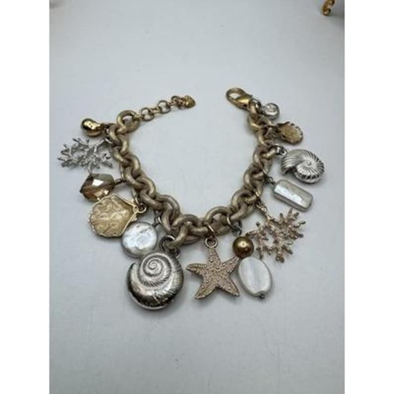 May include: A gold-toned charm bracelet featuring various sea-themed charms. The bracelet includes charms shaped like seashells, starfish, and coral, along with pearl and crystal accents. The chain is a chunky link design with a lobster clasp.