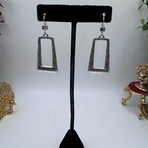 May include: Silver-toned rectangular earrings with a geometric design. The earrings are displayed on a black jewelry stand. The earrings have a rectangular shape with a slightly tapered bottom and a decorative pattern within the frame.