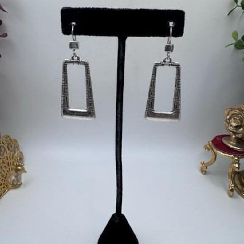 May include: Silver-toned rectangular earrings with a geometric design. The earrings are displayed on a black jewelry stand. The earrings have a rectangular shape with a slightly tapered bottom and a decorative pattern within the frame.