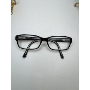 Puede incluir: Gafas rectangulares negras con lentes transparentes. Las monturas son negras con un acento plateado en las patillas. Las gafas est&aacute;n sobre un fondo blanco.