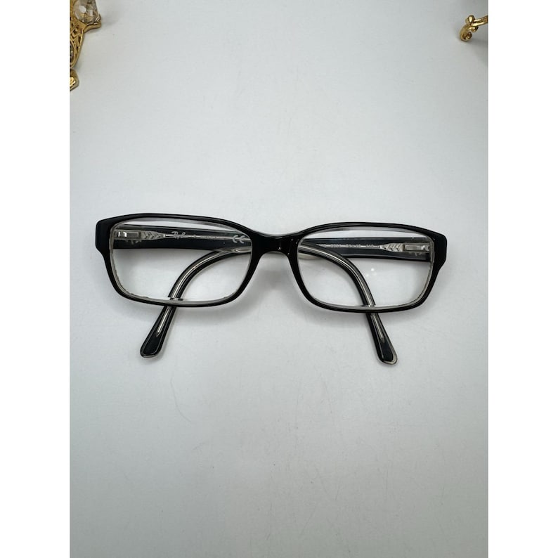 Puede incluir: Gafas rectangulares negras con lentes transparentes. Las monturas son negras con un acento plateado en las patillas. Las gafas est&aacute;n sobre un fondo blanco.