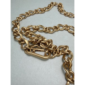 Puede incluir: Una cadena dorada con un dise&ntilde;o tejido y texturizado. La cadena presenta un cierre grande y es adecuada para usar como collar o elemento decorativo. La cadena es de metal y tiene una est&eacute;tica vintage.