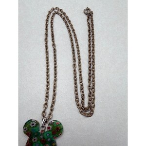 May include: A silver-toned necklace with a pendant shaped like a mouse head. The pendant is green with a floral pattern in red, white, and yellow. The chain is made of small, linked metal pieces.