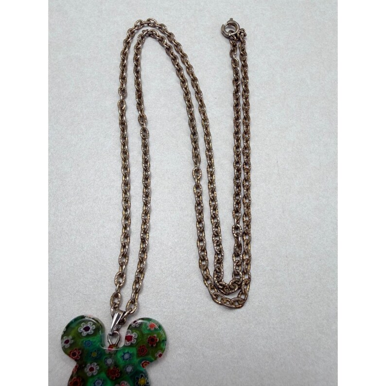 May include: A silver-toned necklace with a pendant shaped like a mouse head. The pendant is green with a floral pattern in red, white, and yellow. The chain is made of small, linked metal pieces.