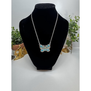 May include: A silver-toned necklace with a butterfly pendant. The butterfly is adorned with turquoise and light green stones. The necklace is displayed on a black mannequin.