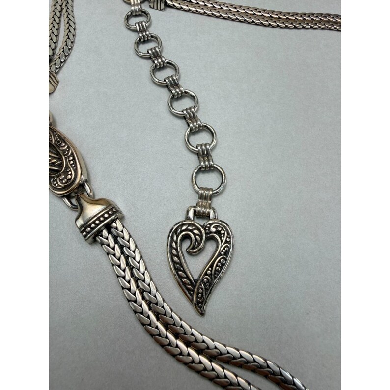 May include: A silver-toned belt with a heart-shaped pendant. The belt features a woven chain design and decorative detailing on the pendant and clasp. The heart pendant is approximately 2 inches in length.