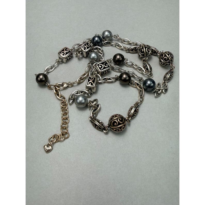 Może przedstawiać: Srebrny naszyjnik z mieszanką ciemnoszarych i srebrnych koralik&oacute;w, a także dekoracyjnych kwadratowych i okrągłych element&oacute;w filigranowych. Naszyjnik posiada łańcuszek z regulowaną długością.