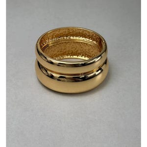 May include: Two gold-colored, hinged bangle bracelets. The bracelets are stacked on top of each other. The top bracelet has a textured interior, while the bottom bracelet has a smooth, polished surface. The bracelets are made of a shiny, metallic material.