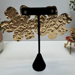 May include: Gold-toned, hammered metal flower earrings displayed on a black velvet earring stand. The earrings have a textured surface and a floral design. The stand has a black cone-shaped base and a vertical black post.