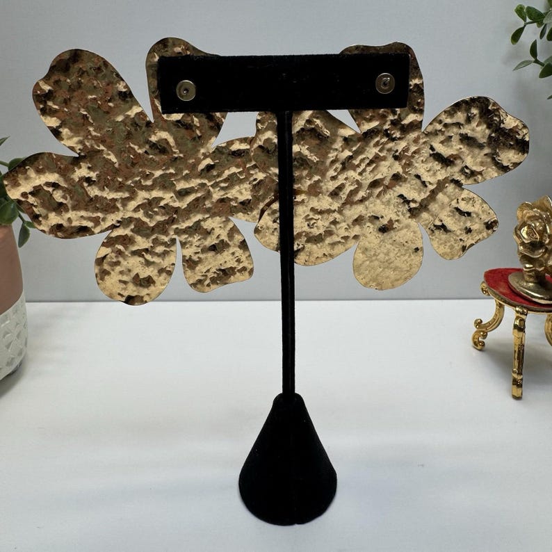 May include: Gold-toned, hammered metal flower earrings displayed on a black velvet earring stand. The earrings have a textured surface and a floral design. The stand has a black cone-shaped base and a vertical black post.