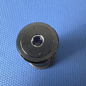 May include: A black, cylindrical object with a circular top featuring a central, dark-colored lens. The top has three small holes and a silver rim. The object is set against a blue textured background.