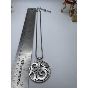 以下が含まれることがあります： 渦巻き模様のデザインが施された、円形のペンダントが付いたシルバーカラーのネックレス。ペンダントの直径は約6.35cmです。サイズの参考として、ネックレスは定規の隣に表示されています。