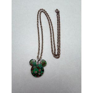 May include: A pendant necklace featuring a Mickey Mouse-shaped charm. The charm is a mosaic of green, red, and white floral patterns. The chain is a delicate, antique bronze color. The necklace is displayed on a light gray surface.