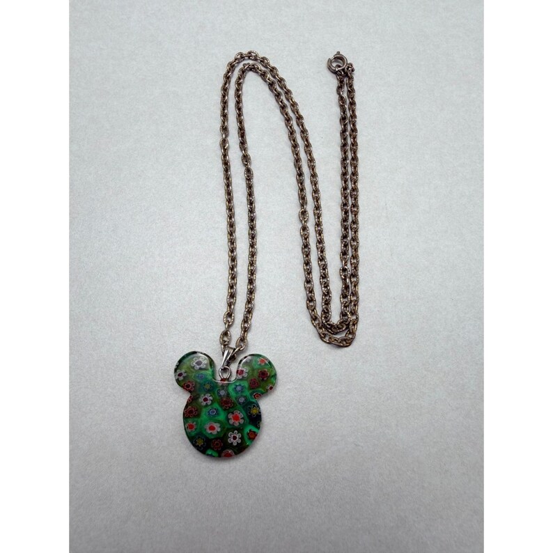 May include: A pendant necklace featuring a Mickey Mouse-shaped charm. The charm is a mosaic of green, red, and white floral patterns. The chain is a delicate, antique bronze color. The necklace is displayed on a light gray surface.