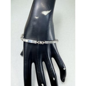 K&ouml;nnte beinhalten: Ein silbernes Armband mit einem rechteckigen Mittelteil und aufwendigen Details. Das Armband hat einen kleinen, runden, klaren Stein und ge&auml;tzte Muster entlang seiner L&auml;nge. Das Armband wird auf einer schwarzen Hand pr&auml;sentiert.