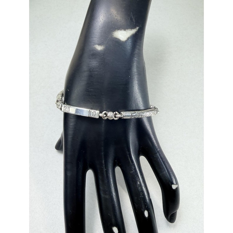 K&ouml;nnte beinhalten: Ein silbernes Armband mit einem rechteckigen Mittelteil und aufwendigen Details. Das Armband hat einen kleinen, runden, klaren Stein und ge&auml;tzte Muster entlang seiner L&auml;nge. Das Armband wird auf einer schwarzen Hand pr&auml;sentiert.