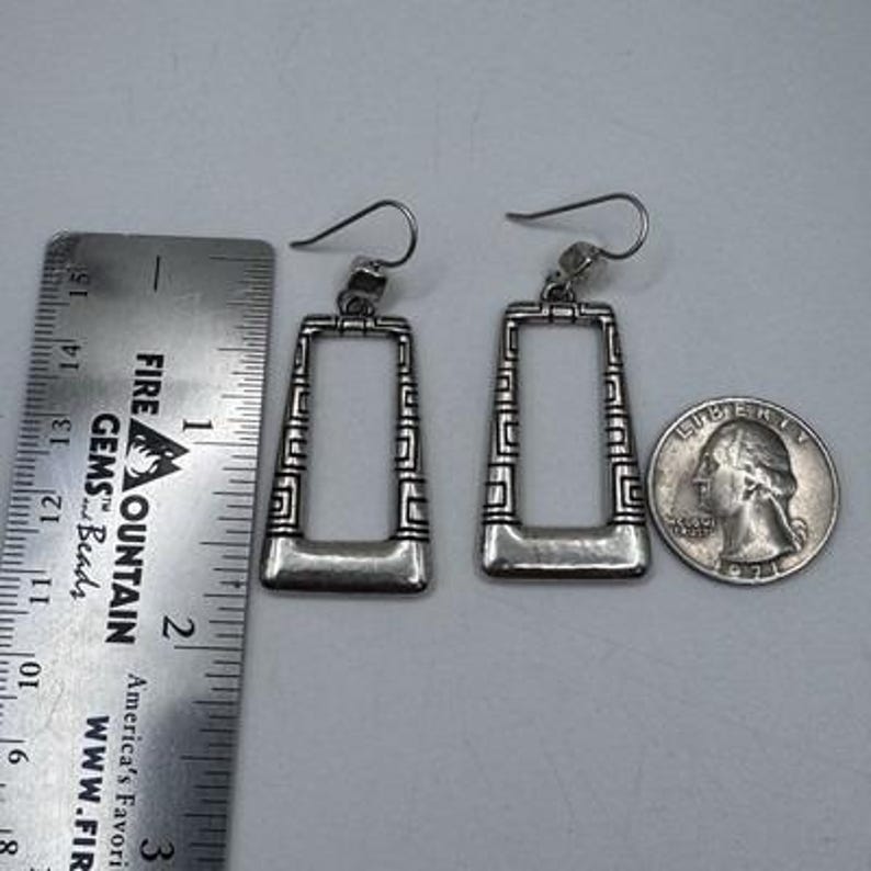May include: Silver-toned rectangular earrings with a geometric design. The earrings are approximately 1.5 inches long. A ruler and a quarter are next to the earrings for size comparison. The earrings have a hook closure.