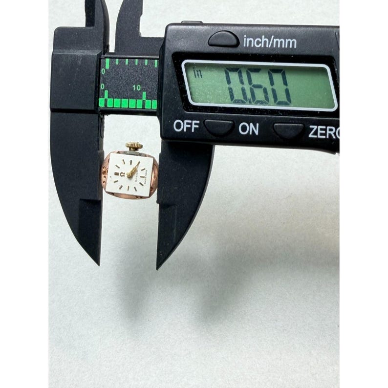 May include: A small, vintage gold-toned watch with a square face, held by a black caliper. The watch face is white with black numerals and hands. The caliper displays a measurement of 1.52 cm.