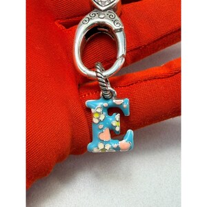 Peut inclure: Un charm argent&eacute; avec la lettre "E" en &eacute;mail bleu, orn&eacute; de motifs floraux blancs et roses. Le charm est attach&eacute; &agrave; un fermoir argent&eacute; avec un motif de c&oelig;ur. Le fond est en tissu rouge.