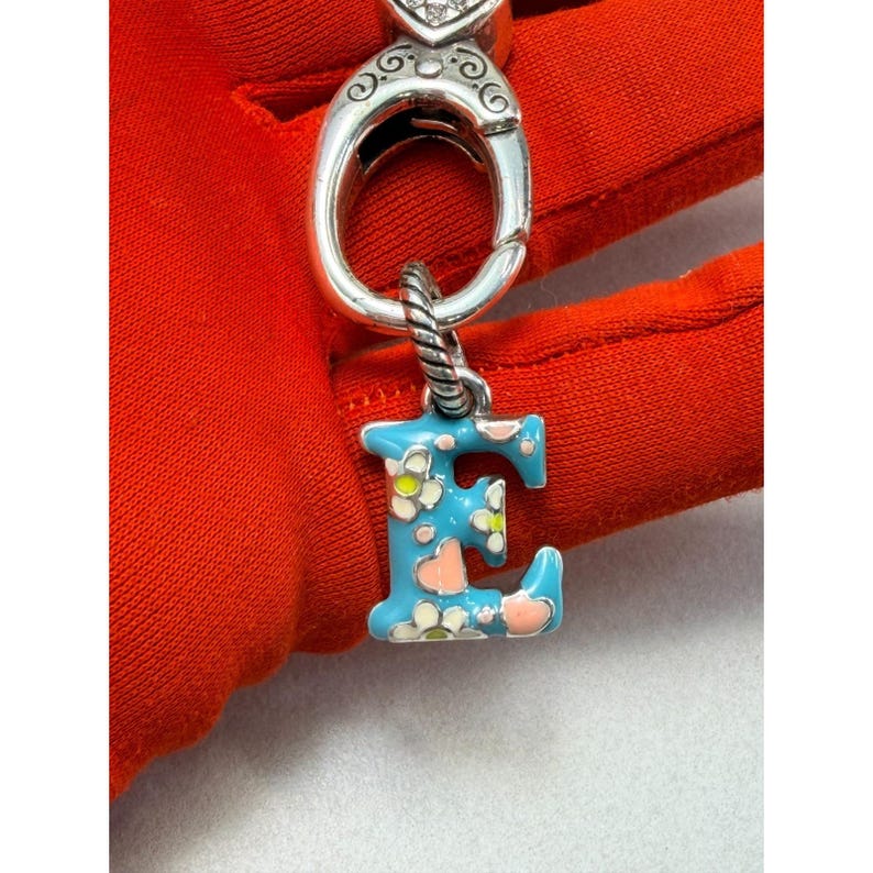 Peut inclure: Un charm argent&eacute; avec la lettre "E" en &eacute;mail bleu, orn&eacute; de motifs floraux blancs et roses. Le charm est attach&eacute; &agrave; un fermoir argent&eacute; avec un motif de c&oelig;ur. Le fond est en tissu rouge.
