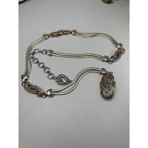 May include: Silver-toned metal belt with ornate, swirling designs and a decorative pendant. The belt features a chain-link style and a series of decorative clasps. The belt is adjustable with a chain and hook closure.