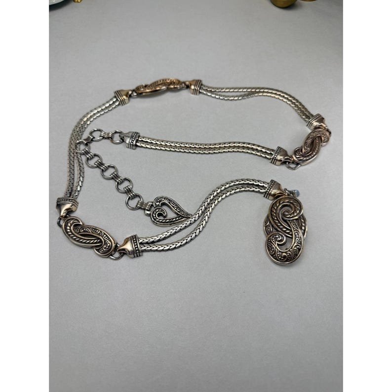 May include: Silver-toned metal belt with ornate, swirling designs and a decorative pendant. The belt features a chain-link style and a series of decorative clasps. The belt is adjustable with a chain and hook closure.