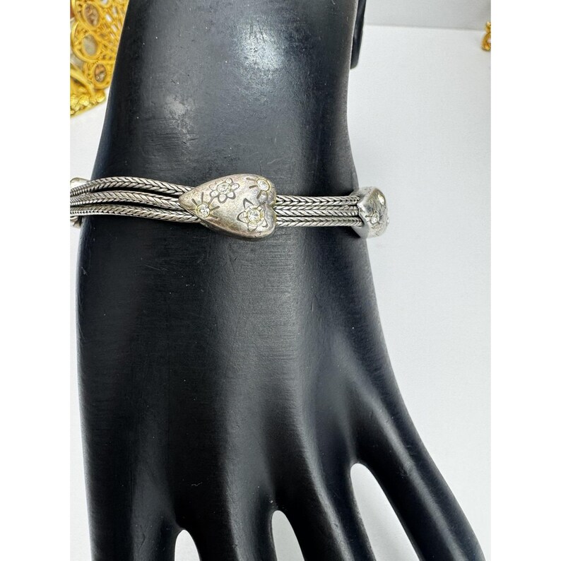 May include: Silver bracelet with a heart-shaped central charm decorated with flowers and small crystals. The bracelet has a braided design and a small, round charm at the end. The jewellery is displayed on a black hand-shaped mannequin.