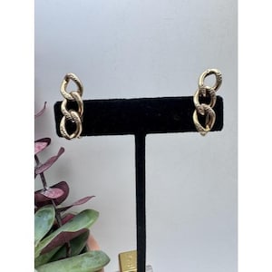 Op de afbeelding: Een paar goudkleurige oorringen met schakelketting. Elke oorring heeft drie in elkaar grijpende ovale schakels met een textuurpatroon. De oorbellen worden op een zwarte standaard tegen een witte achtergrond gepresenteerd.