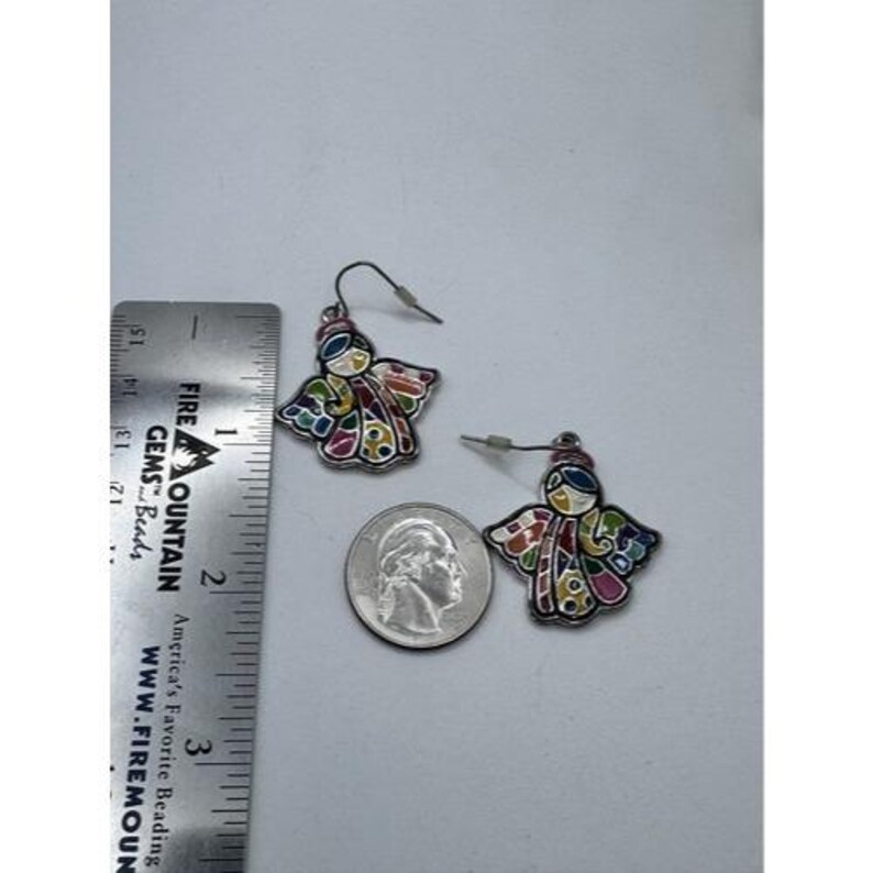 May include: A pair of angel-shaped earrings with colorful enamel wings and a silver-tone metal outline. The earrings are shown next to a ruler and a quarter for size reference. The earrings are approximately 1 inch in length.