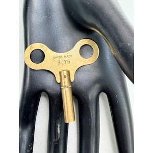 May include: A vintage brass clock key with two circular openings and a cylindrical winding post. The key is stamped with "SWISS MADE" and "3.75". The key is a golden color.