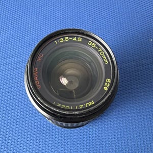 May include: A black Osawa MC 35-70mm camera lens with a 52mm diameter. The lens has the specifications 1:3.5-4.5 printed on the top and the serial number 1770127 on the bottom. The lens is set against a blue background.