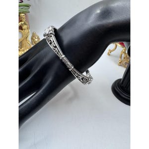 May include: A silver-toned bracelet with ornate scrollwork and a square, clear stone. The bracelet is displayed on a black hand-shaped jewelry holder, showcasing its intricate design and craftsmanship. The bracelet's design is symmetrical.