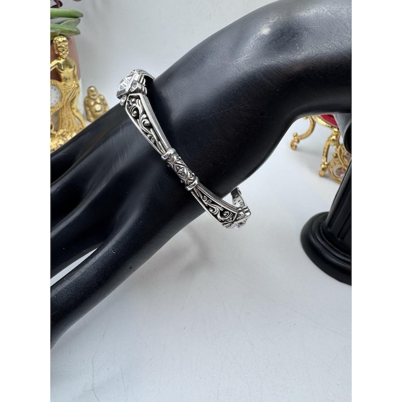 May include: A silver-toned bracelet with ornate scrollwork and a square, clear stone. The bracelet is displayed on a black hand-shaped jewelry holder, showcasing its intricate design and craftsmanship. The bracelet's design is symmetrical.