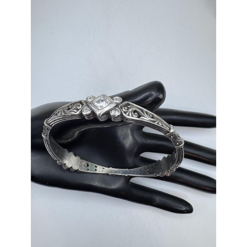 May include: Silver-toned hinged bracelet with a central square clear stone and smaller round stones. The bracelet features ornate scrollwork and a decorative design. The bracelet is shown on a black surface.