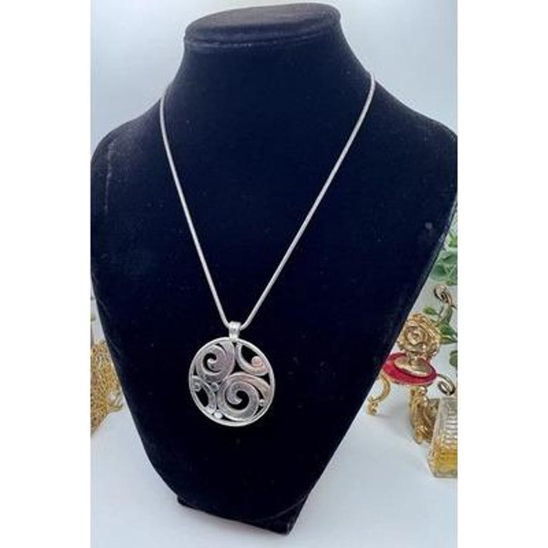 以下が含まれることがあります： シルバーカラーのペンダントネックレス。ペンダントは渦巻き模様の円形デザインです。ネックレスは黒いマネキンに展示されています。背景には小さな装飾椅子やその他のアイテムが含まれています。