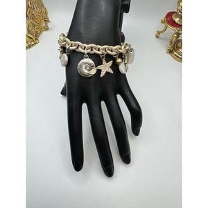 May include: A silver-toned charm bracelet with a chunky chain design. The bracelet features various charms, including a seashell, a starfish, and other decorative elements. The bracelet is displayed on a black hand.