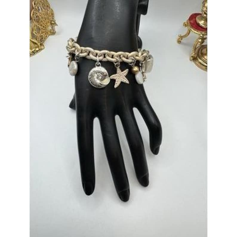 May include: A silver-toned charm bracelet with a chunky chain design. The bracelet features various charms, including a seashell, a starfish, and other decorative elements. The bracelet is displayed on a black hand.