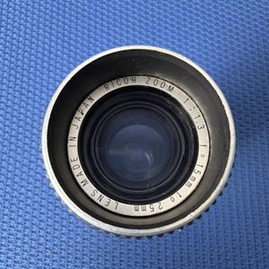 May include: A close-up view of a Ricoh Zoom camera lens. The lens is silver with black accents and features the text "Made in Japan". The lens has a focal length of 15mm to 25mm and an aperture of 1:1.3.