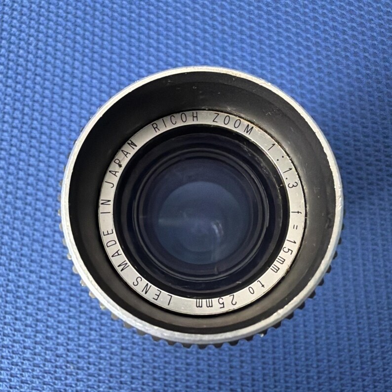 May include: A close-up view of a Ricoh Zoom camera lens. The lens is silver with black accents and features the text "Made in Japan". The lens has a focal length of 15mm to 25mm and an aperture of 1:1.3.