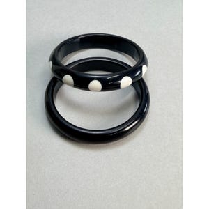 May include: Two circular bangle bracelets with a smooth, glossy finish. One bangle is solid black, while the other is black with a repeating pattern of white circular polka dots spaced evenly around the outer surface of the jewelry piece.