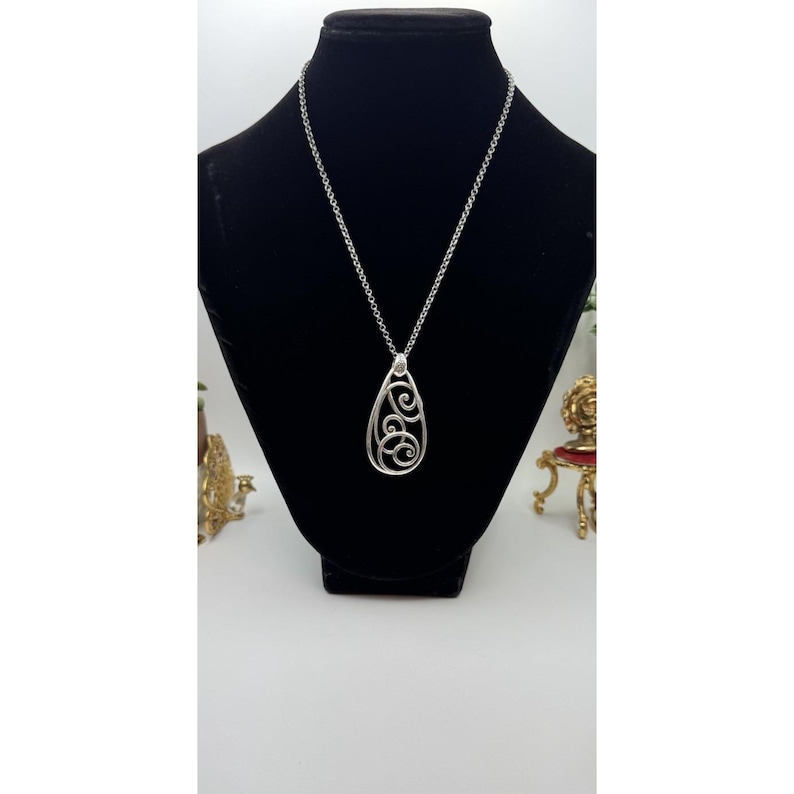 May include: A silver-colored pendant necklace with a teardrop-shaped pendant. The pendant features an openwork design of swirling patterns. The necklace is displayed on a black mannequin, showcasing the jewelry's design and details.