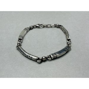 K&ouml;nnte beinhalten: Ein silbernes Armband mit einem verzierten Design. Das Armband hat eine Reihe von abgerundeten Perlen und rechteckigen Segmenten mit ge&auml;tzten Mustern. Der Verschluss ist ein einfacher Haken und eine Kette. Das Armband wird auf grauem Hintergrund gezeigt.