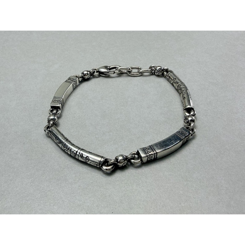 K&ouml;nnte beinhalten: Ein silbernes Armband mit einem verzierten Design. Das Armband hat eine Reihe von abgerundeten Perlen und rechteckigen Segmenten mit ge&auml;tzten Mustern. Der Verschluss ist ein einfacher Haken und eine Kette. Das Armband wird auf grauem Hintergrund gezeigt.