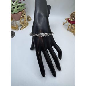 May include: Silver-toned bracelet with an ornate design. The bracelet features a central square-shaped clear stone, flanked by smaller clear stones. The bracelet is displayed on a black hand-shaped jewelry display.