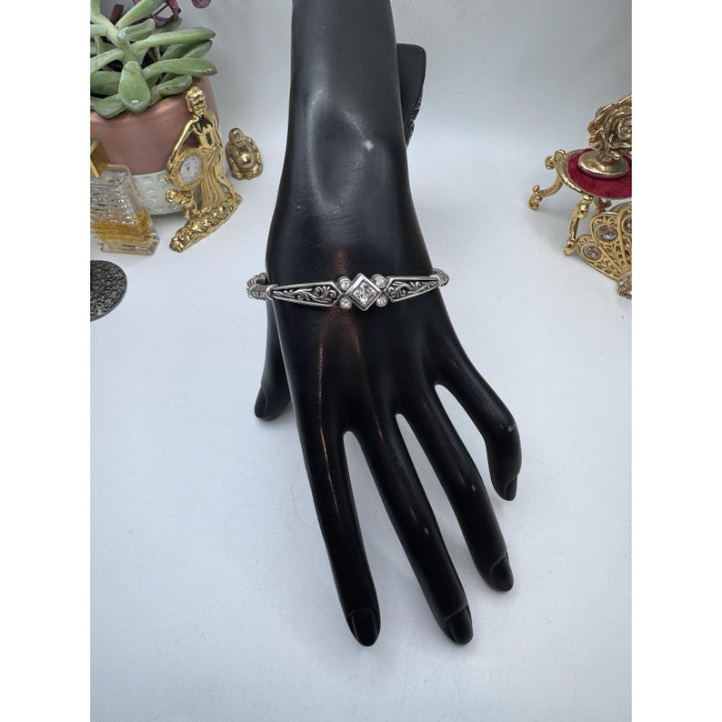 May include: Silver-toned bracelet with an ornate design. The bracelet features a central square-shaped clear stone, flanked by smaller clear stones. The bracelet is displayed on a black hand-shaped jewelry display.