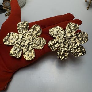May include: Two gold-colored flower-shaped earrings with a textured surface. The earrings are displayed on a red surface. The earrings have a hammered metal appearance and are likely costume jewelry.