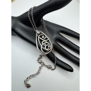 May include: Silver-toned pendant necklace with an open teardrop design. The pendant features an intricate, swirling pattern within the frame. The necklace has a delicate chain with heart-shaped accents near the clasp and a heart-shaped charm at the end.